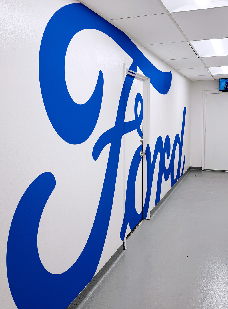 White hallway wall painted with a large, bold blue Ford script logo spanning across doors and extending down the corridor.