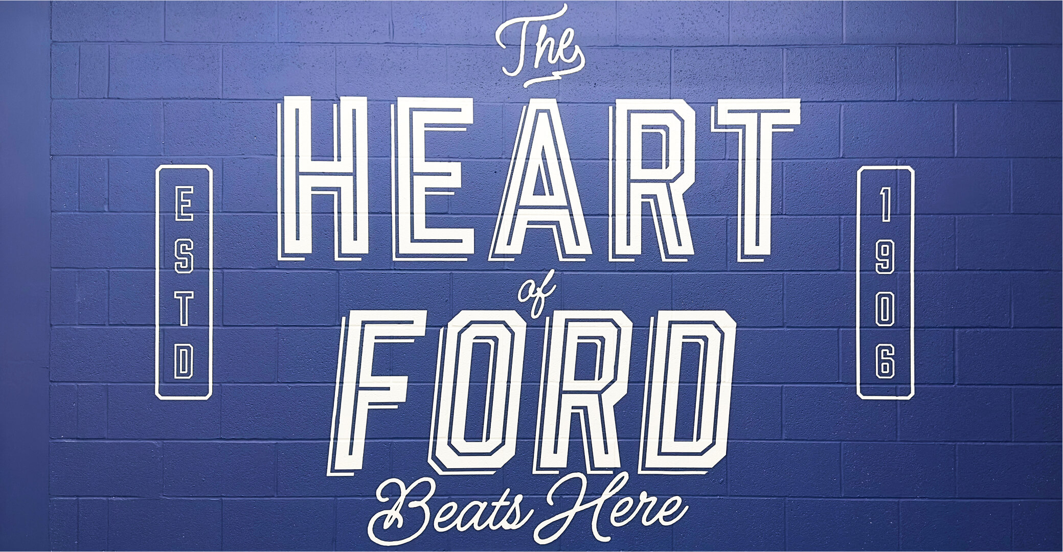 Large white lettering on a blue brick wall reads 'The Heart of Ford Beats Here' with 'ESTD 1906' displayed vertically on each side.