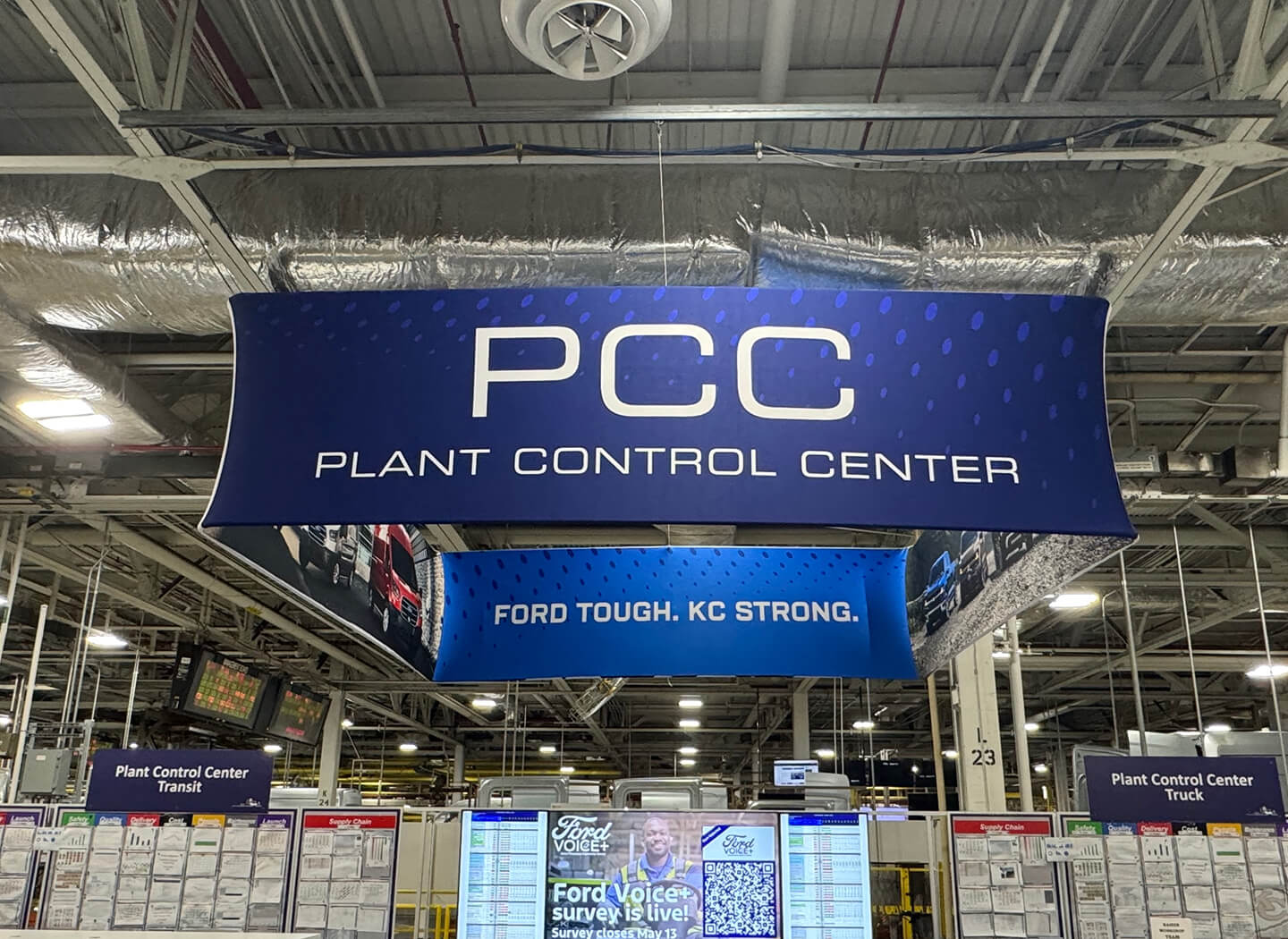 Overhead blue banner with white text reading 'PCC Plant Control Center' and 'Ford Tough. KC Strong.' hanging above workstations and display boards.