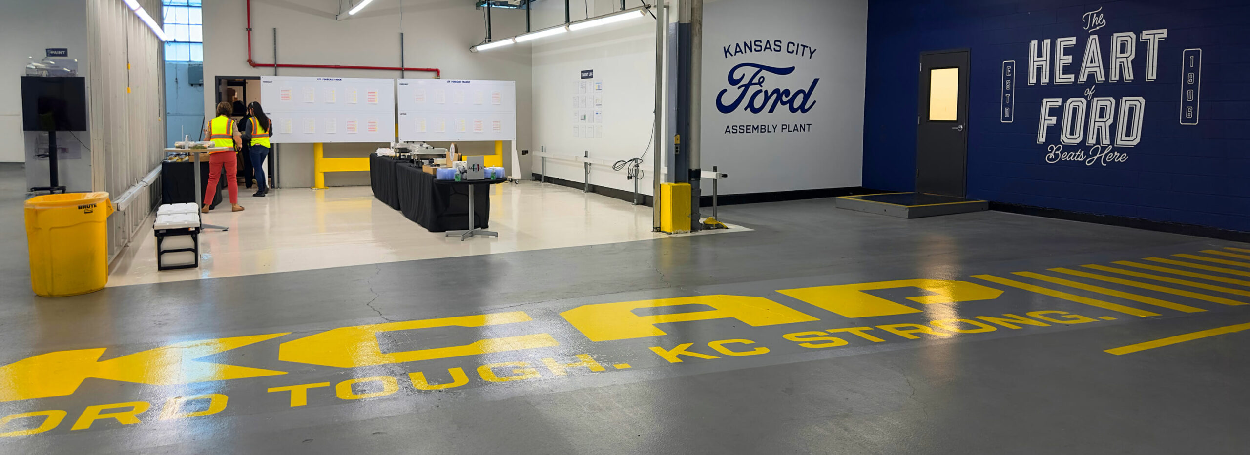 Factory space with Kansas City Ford Assembly Plant and 'The Heart of Ford Beats Here' wall graphics, yellow KCAP floor lettering, and workers in safety vests near tables and boards.