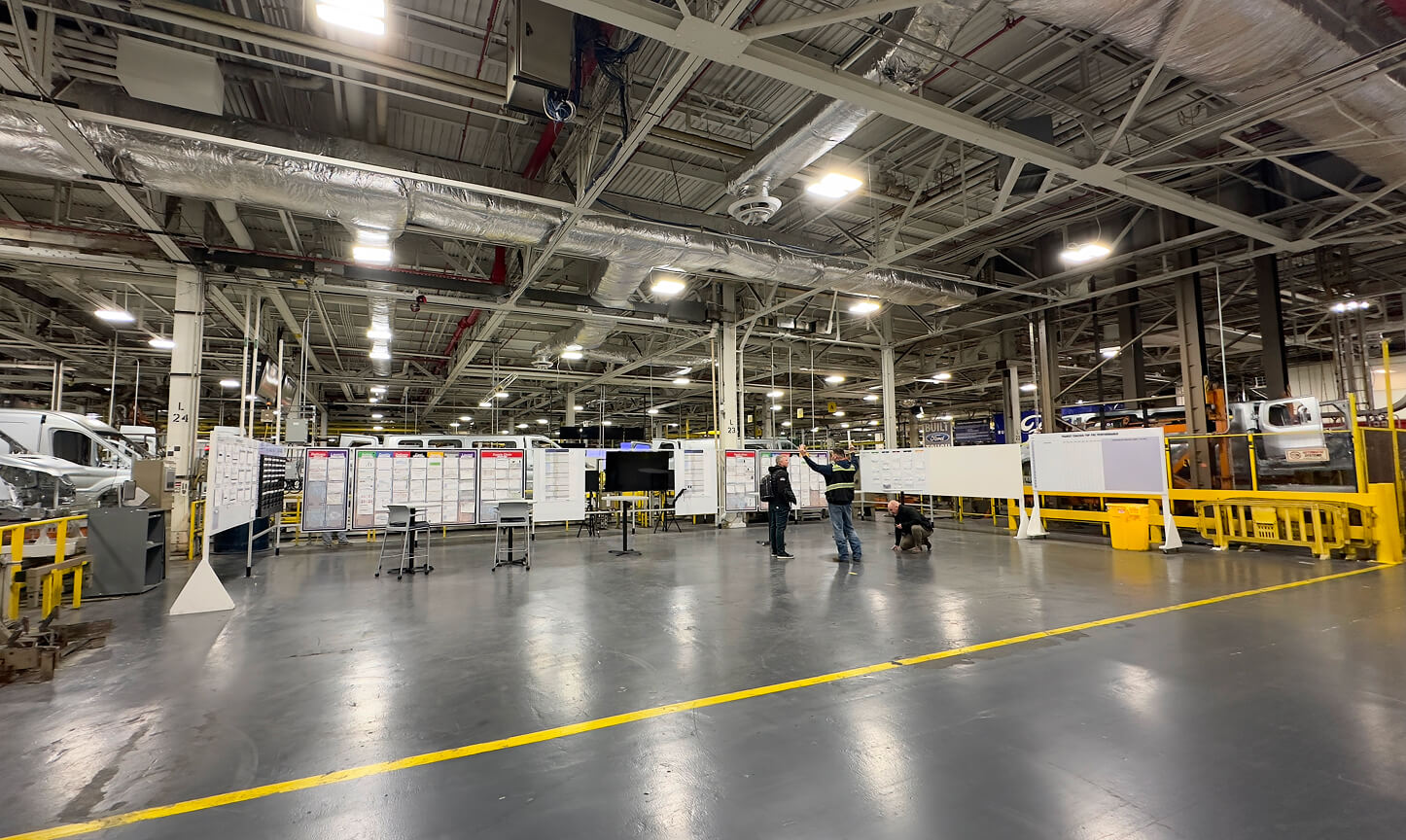 Large industrial warehouse interior with bright overhead lighting, whiteboards, and a few workers standing and crouching on the polished concrete floor.