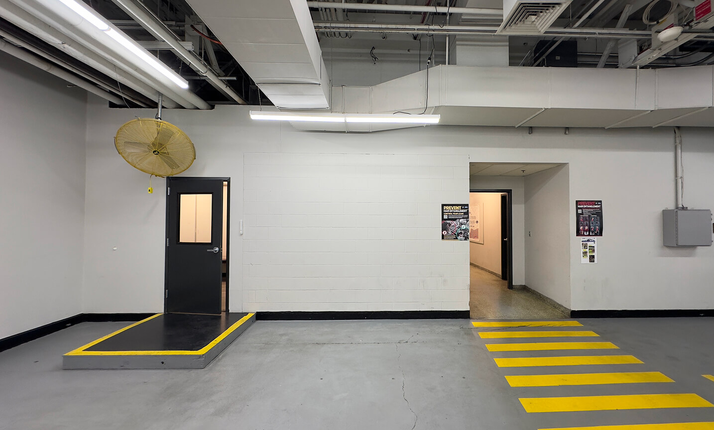 Gray-walled interior with overhead pipes, a yellow safety fan, two doors, and bold yellow-striped walkway markings on the concrete floor.