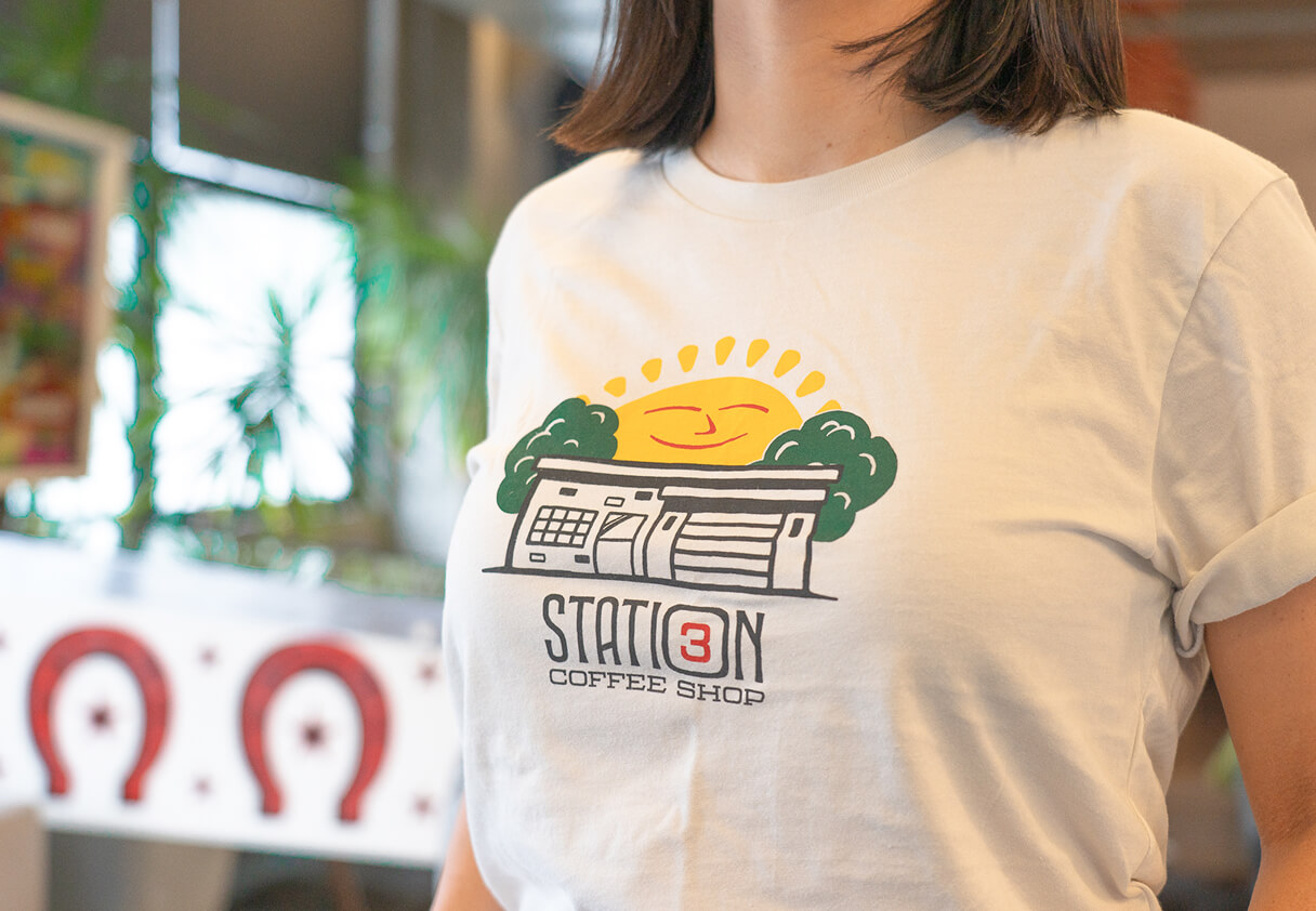 Person wearing a white t-shirt featuring the Station 3 Coffee Shop logo with a smiling sunrise, trees, and building illustration.