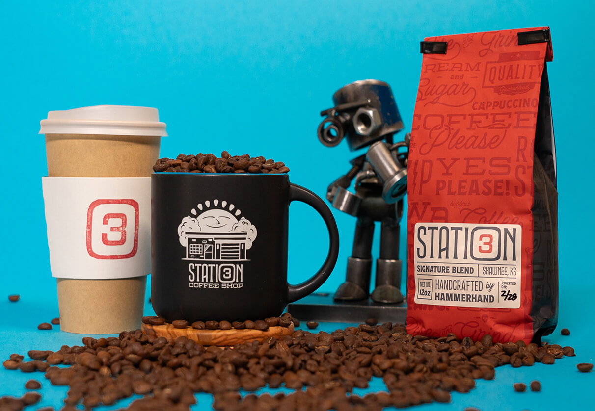 Coffee bag, branded black mug filled with beans, takeaway cup with logo sleeve, and small metal robot figure against blue background.