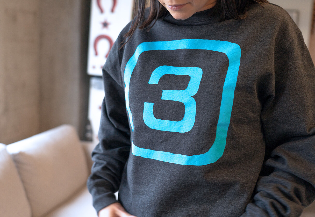 Person wearing a dark gray sweatshirt with a large turquoise Station 3 logo featuring a bold number 3 inside a rounded square.